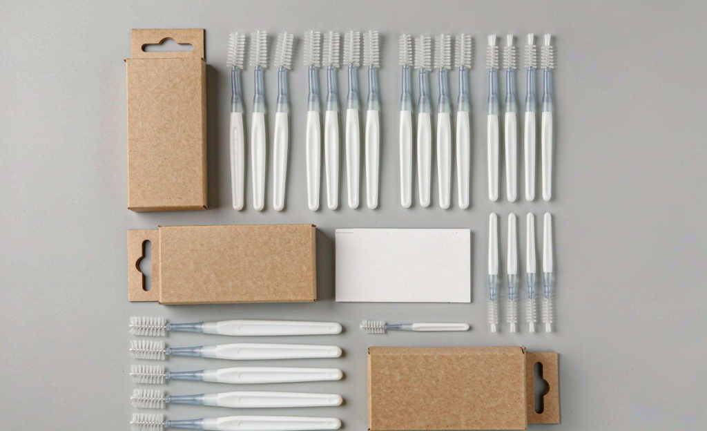 Flat-lay of plain white interdental brushes in assorted sizes with unbranded kraft cartons on a light grey studio background